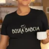 Zbliżenie na czarną koszulkę z napisem Boska Babcia i imieniem Marysia w sercu, modelka trzyma biały kubek