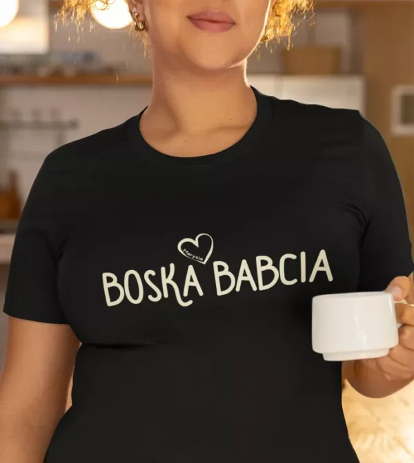Zbliżenie na czarną koszulkę z napisem Boska Babcia i imieniem Marysia w sercu, modelka trzyma biały kubek