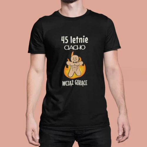 Mężczyzna w czarnej koszulce z nadrukiem „45-letnie CIACHO WCIĄŻ GORĄCE”. Grafika przedstawia uśmiechniętego, umięśnionego piernika z muszką, otoczonego płomieniami. Koszulka urodzinowa z humorem, idealna na prezent dla mężczyzny.