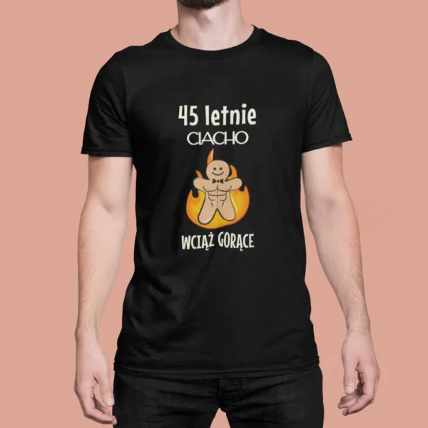 Mężczyzna w czarnej koszulce z nadrukiem „45-letnie CIACHO WCIĄŻ GORĄCE”. Grafika przedstawia uśmiechniętego, umięśnionego piernika z muszką, otoczonego płomieniami. Koszulka urodzinowa z humorem, idealna na prezent dla mężczyzny.