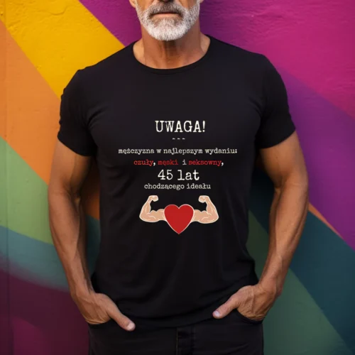 Mężczyzna w czarnej koszulce z nadrukiem "Uwaga! Mężczyzna w najlepszym wydaniu: czuły, męski i seksowny, 45 lat chodzącego ideału". Grafika zawiera także dwa umięśnione ramiona i czerwone serce. Tło kolorowe, geometryczne. Koszulka przeznaczona na prezent urodzinowy.