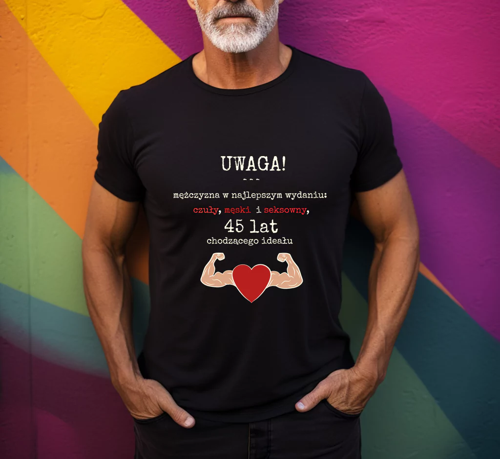 Mężczyzna w czarnej koszulce z nadrukiem "Uwaga! Mężczyzna w najlepszym wydaniu: czuły, męski i seksowny, 45 lat chodzącego ideału". Grafika zawiera także dwa umięśnione ramiona i czerwone serce. Tło kolorowe, geometryczne. Koszulka przeznaczona na prezent urodzinowy.