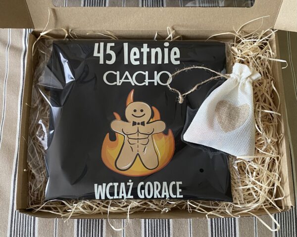 szare kartonowe pudełeczko wypełnione siankiem z koszulką 45 letnie ciacho i woreczkiem z serduszkiem zawierające odrobinę słodkości. Zestaw prezentowy z koszulką na urodziny 45 letnie ciacho - wciaz gorące