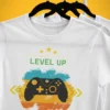 Biała koszulka dla gracza Level Up! z kontrolerem i napisem „Unlocked Level 19”, wisząca na wieszaku.