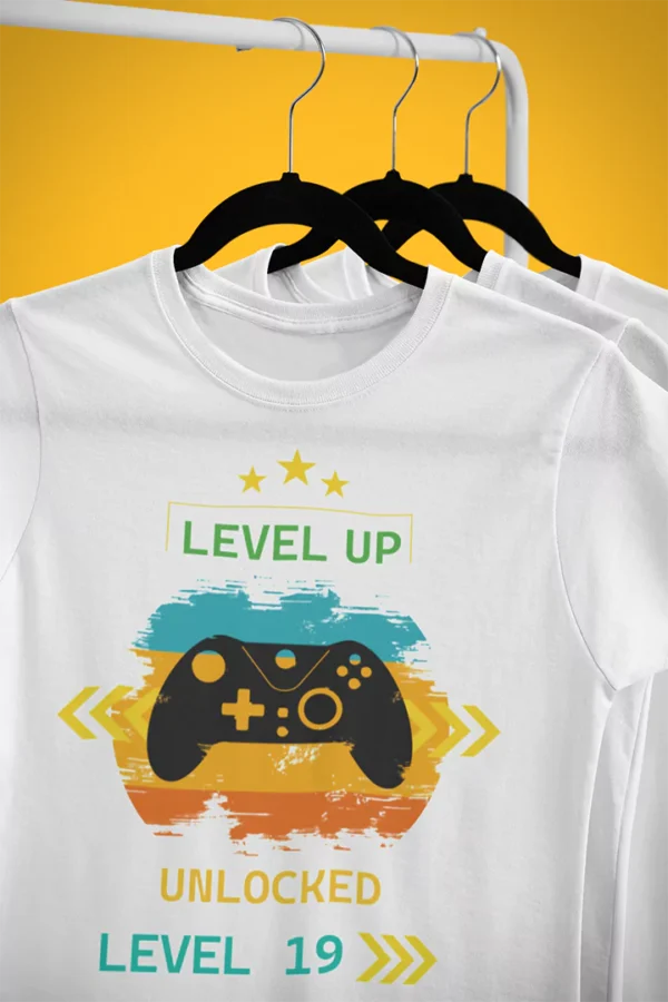 Biała koszulka dla gracza Level Up! z kontrolerem i napisem „Unlocked Level 19”, wisząca na wieszaku.