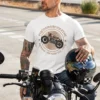 Mężczyzna w białej koszulce 'Pięćdziesięcioletni Klasyk' z motocyklem, siedzący na motorze, trzymający kask.