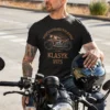 Mężczyzna w czarnej koszulce 'Pięćdziesięcioletni Klasyk' z motocyklem, siedzący na motorze, trzymający kask.