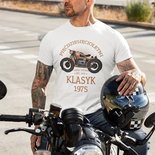 Mężczyzna w białej koszulce 'Pięćdziesięcioletni Klasyk EST. 1975' z tatuażami, oparty o motocykl, z kaskiem na tle ulicy.