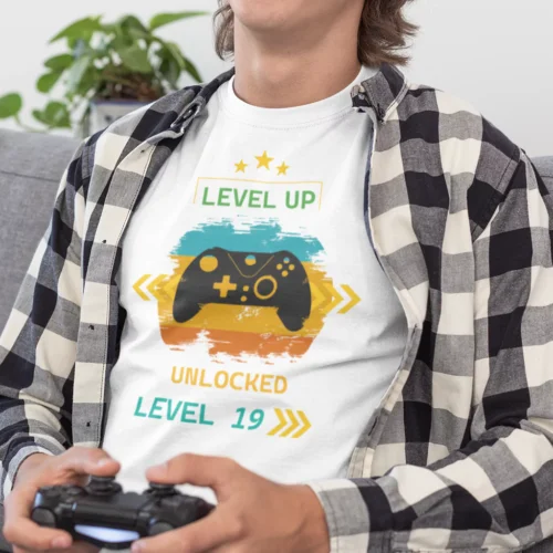 Mężczyzna w białej koszulce dla gracza Level Up! z kontrolerem, uśmiechnięty, grający na konsoli siedząc na szarej kanapie.