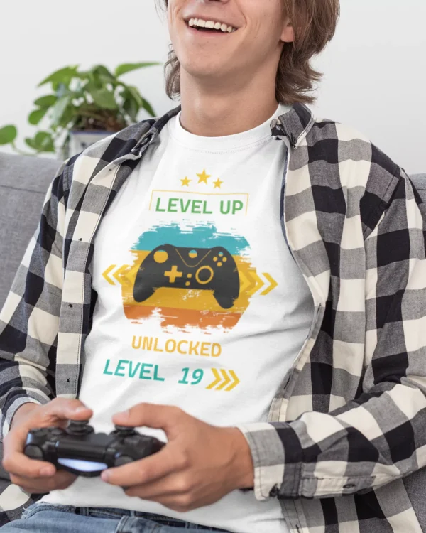 Mężczyzna w białej koszulce dla gracza Level Up! z kontrolerem, uśmiechnięty, grający na konsoli siedząc na szarej kanapie.