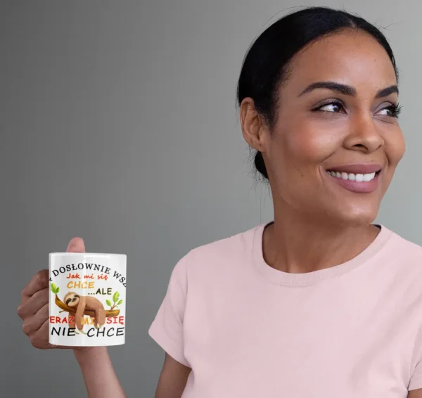 Uśmiechnięta kobieta w różowej koszulce trzymająca biały kubek z nadrukiem leniwca i napisem "Teraz mi się nie chce", patrząca w bok, na szarym tle.