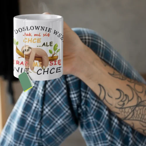 Osoba z tatuażami na rękach, w piżamowych spodniach w kratę, trzymająca biały kubek z parującą herbatą i torebką herbaty, z nadrukiem leniwca i napisem "Teraz mi się nie chce".