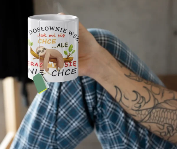 Osoba z tatuażami na rękach, w piżamowych spodniach w kratę, trzymająca biały kubek z parującą herbatą i torebką herbaty, z nadrukiem leniwca i napisem "Teraz mi się nie chce".