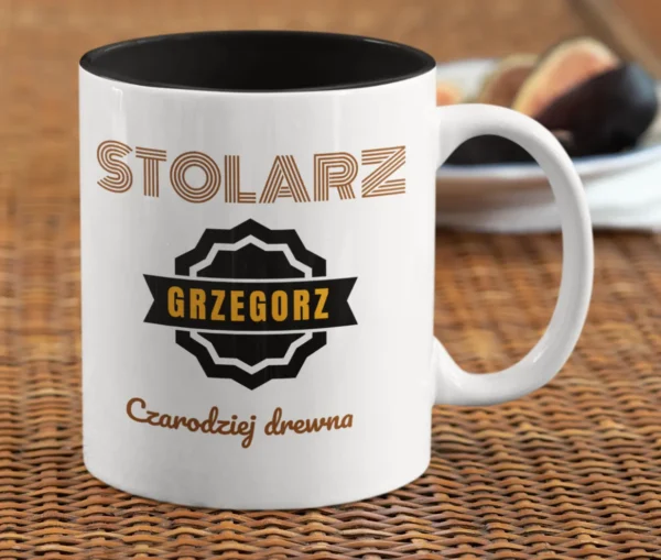 Personalizowany kubek z czarnym wnętrzem, napisem 'STOLARZ GRZEGORZ Czarodziej drewna' i figami w tle, na rattanowej podkładce.
