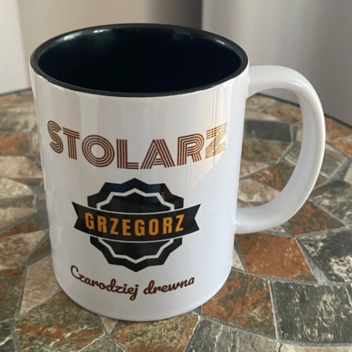 Biały kubek z czarnym środkiem i personalizowanym nadrukiem 'STOLARZ GRZEGORZ Czarodziej drewna', stojący na mozaikowym blacie.