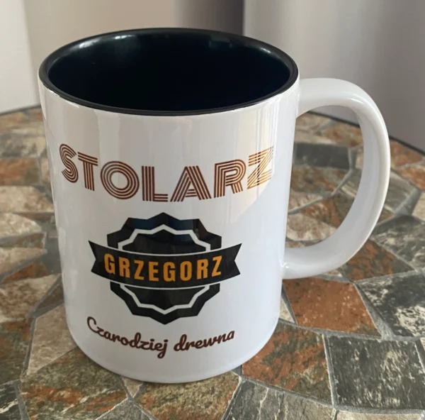 Biały kubek z czarnym środkiem i personalizowanym nadrukiem 'STOLARZ GRZEGORZ Czarodziej drewna', stojący na mozaikowym blacie.