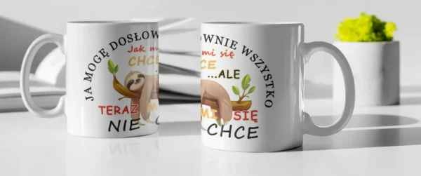 Dwa białe ceramiczne kubki z nadrukiem leniwca i cytatem "Teraz mi się nie chce" stojące obok siebie na białym stole z rozmazanym tłem książek i zielonej dekoracji.