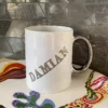 Biały personalizowany kubek ceramiczny ze srebrnym uchem i srebrnym napisem "DAMIAN", stojący na kolorowej tkaninie.