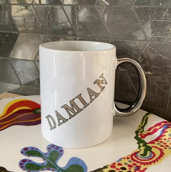 Biały personalizowany kubek ceramiczny ze srebrnym uchem i srebrnym napisem "DAMIAN", stojący na kolorowej tkaninie.