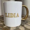 Biały kubek ceramiczny ze złotym uchem i złotym napisem "LIDIA", stojący na mozaikowym stole.