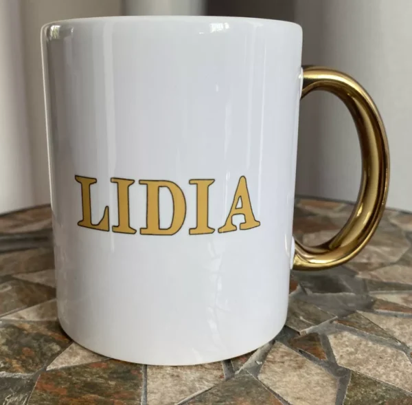 Biały kubek ceramiczny ze złotym uchem i złotym napisem "LIDIA", stojący na mozaikowym stole.