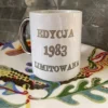 Biały kubek ceramiczny ze srebrnym uchem i srebrnym napisem "EDYCJA 1983 LIMITOWANA", widoczny na tle wzorzystej tkaniny.