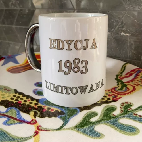 Biały kubek ceramiczny ze srebrnym uchem i srebrnym napisem "EDYCJA 1983 LIMITOWANA", widoczny na tle wzorzystej tkaniny.