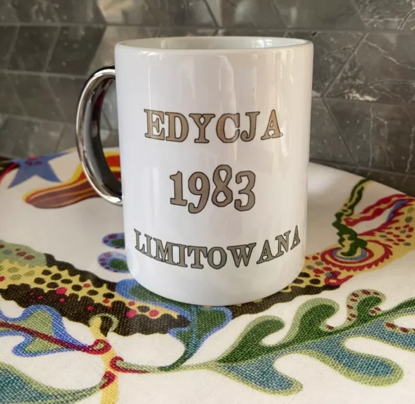 Biały kubek ceramiczny ze srebrnym uchem i srebrnym napisem "EDYCJA 1983 LIMITOWANA", widoczny na tle wzorzystej tkaniny.