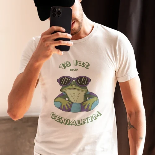 Brodaty mężczyzna z tatuażami robiący selfie, ubrany w białą koszulkę urodzinową z nadrukiem wyluzowanej żaby w okularach i tekstem 'X lat bycia GENIALNYM'.