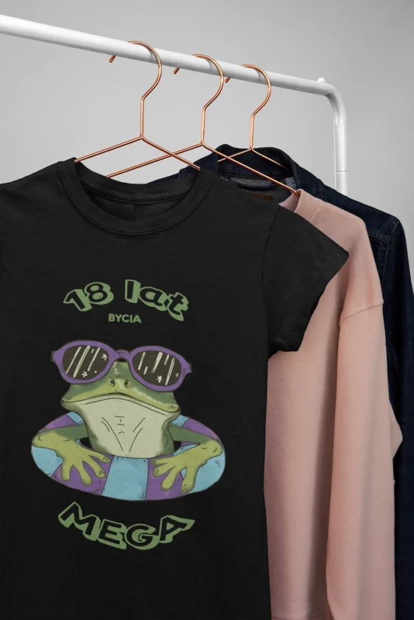 Zbliżenie na nadruk na czarnej koszulce: zielona żaba w fioletowych okularach przeciwsłonecznych, w turkusowo-fioletowym kółku do pływania, z napisem '18 lat bycia mega'