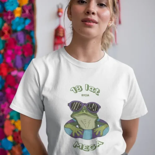 Uśmiechnięta kobieta w białej koszulce z nadrukiem zielonej żaby w okularach i personalizowanym wiekiem, pozująca na tle ściany