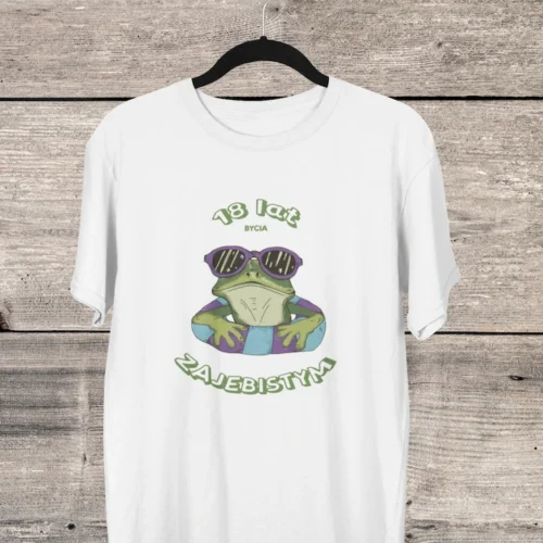 Biała koszulka urodzinowa męska z zabawną żabą w okularach i napisem '18 lat bycia zajebistym', wisząca na drewnianym tle.