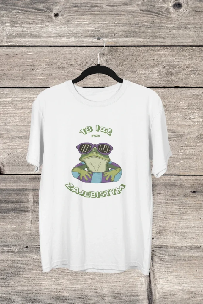 Biała koszulka urodzinowa męska z zabawną żabą w okularach i napisem '18 lat bycia zajebistym', wisząca na drewnianym tle.