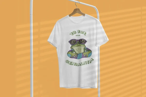 Biała męska koszulka urodzinowa z nadrukiem wesołej żaby w okularach i personalizowanym wiekiem 'X lat bycia GENIALNYM', wisząca na wieszaku.