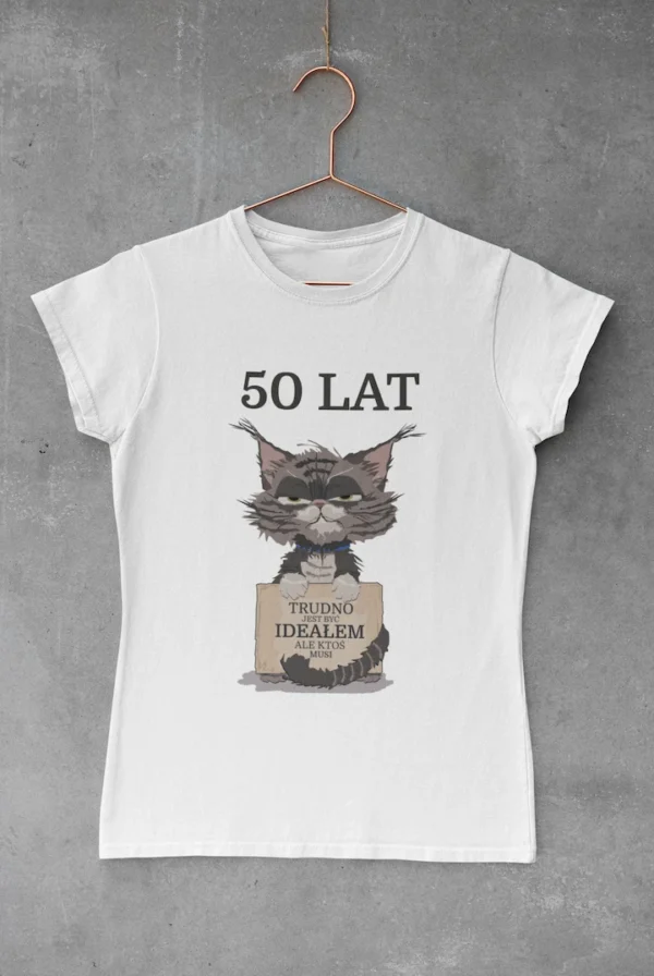 Biała damska koszulka urodzinowa z napisem "Trudno być ideałem" na wieszaku Biała damska koszulka z okrągłym dekoltem, z nadrukiem "50 LAT TRUDNO BYĆ IDEAŁEM ALE KTOŚ MUSI", wisząca na miedzianym wieszaku na tle szarej ściany.