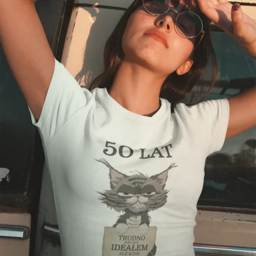 Młoda kobieta w białej koszulce urodzinowej z nadrukiem "50 LAT TRUDNO BYĆ IDEAŁEM ALE KTOŚ MUSI" pozuje przy samochodzie.