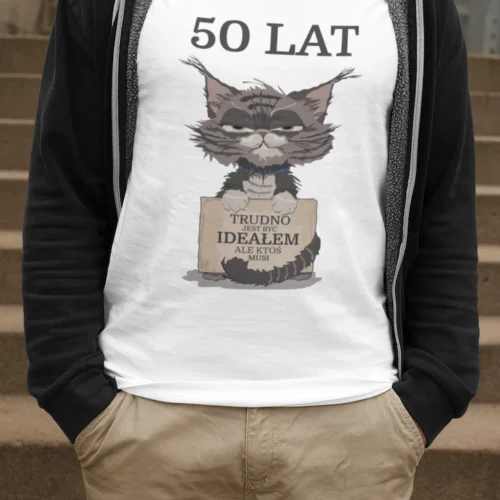 Mężczyzna w białej męskiej koszulce urodzinowej z personalizowanym nadrukiem '50 LAT TRUDNO JEST BYĆ IDEAŁEM ALE KTOŚ MUSI' i grafiką zrzędliwego kota, stojący na schodach.
