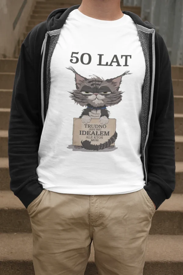 Mężczyzna w białej męskiej koszulce urodzinowej z personalizowanym nadrukiem '50 LAT TRUDNO JEST BYĆ IDEAŁEM ALE KTOŚ MUSI' i grafiką zrzędliwego kota, stojący na schodach.