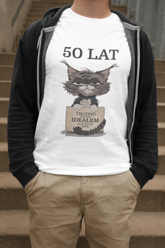 Mężczyzna w białej męskiej koszulce urodzinowej z personalizowanym nadrukiem '50 LAT TRUDNO JEST BYĆ IDEAŁEM ALE KTOŚ MUSI' i grafiką zrzędliwego kota, stojący na schodach.