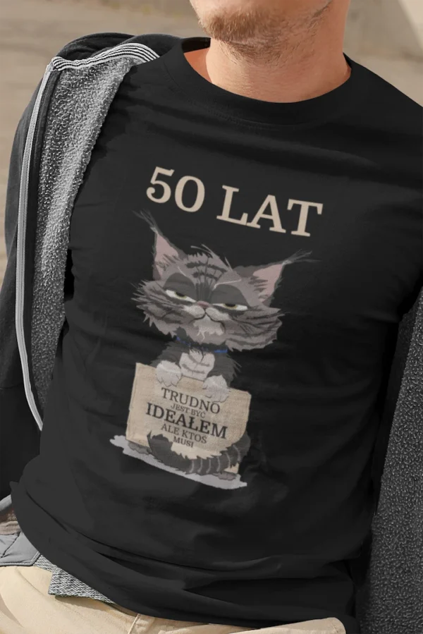 Zbliżenie na czarną męską koszulkę urodzinową z nadrukiem '50 LAT TRUDNO JEST BYĆ IDEAŁEM ALE KTOŚ MUSI' i wizerunkiem zrzędliwego kota, noszoną przez mężczyznę.