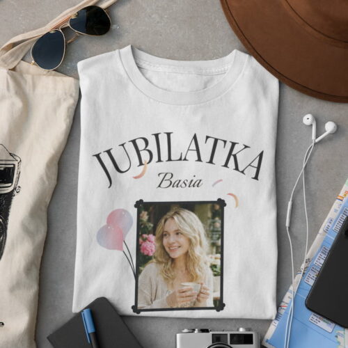 Złożona biała koszulka z personalizacją leżąca obok aparatu fotograficznego, okularów i mapy