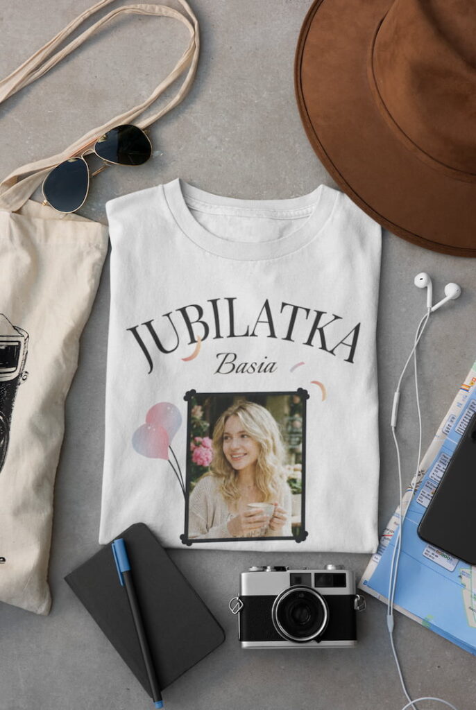 Złożona biała koszulka z personalizacją leżąca obok aparatu fotograficznego, okularów i mapy