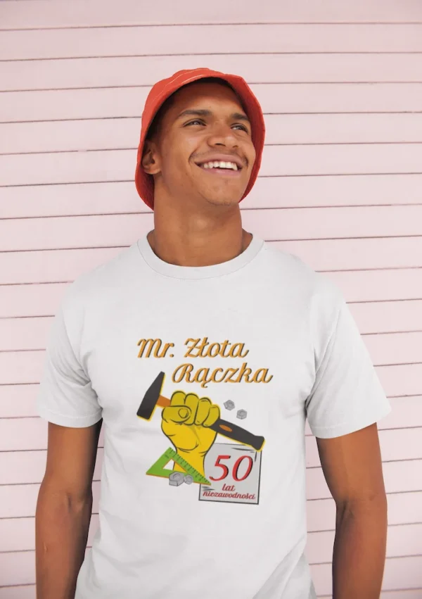 Młody mężczyzna w białej koszulce z nadrukiem Złota Rączka uśmiecha się na tle drewnianej ściany.