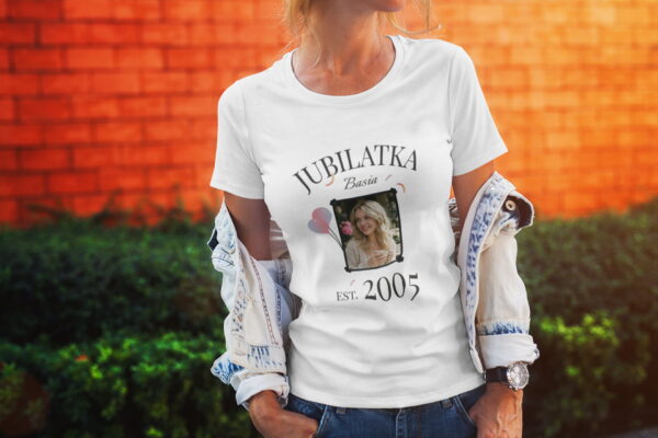Kobieta w białej koszulce z napisem Jubilatka, własnym zdjęciem i datą 2005 na tle ceglanej ściany