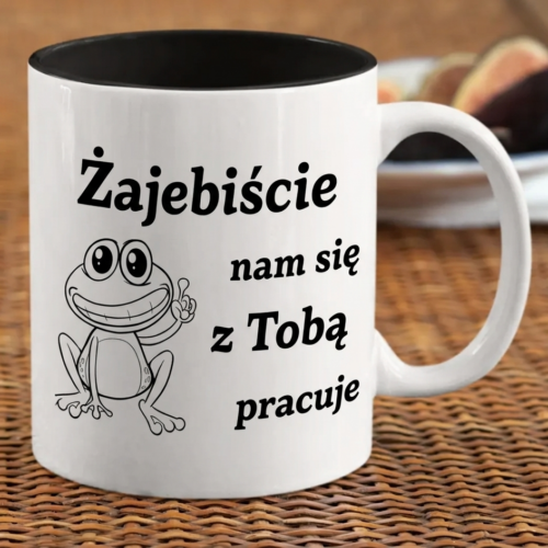 czarny ceramiczny kubek z napisem Zajebiście nam się z Tobą pracuje oraz ilustracją uśmiechniętej żaby na marmurowym blacie.