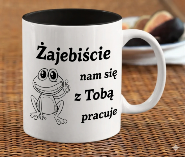 czarny ceramiczny kubek z napisem Zajebiście nam się z Tobą pracuje oraz ilustracją uśmiechniętej żaby na marmurowym blacie.