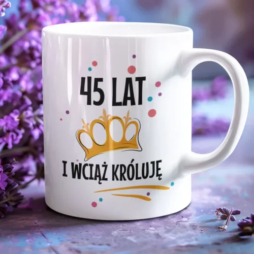 Biały ceramiczny kubek z napisem 45 lat i wciąż króluje oraz grafiką złotej korony na tle kwiatów