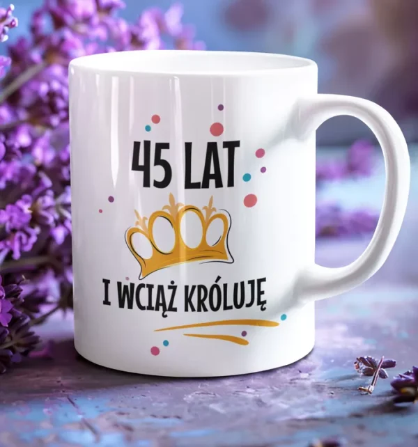 Biały ceramiczny kubek z napisem 45 lat i wciąż króluje oraz grafiką złotej korony na tle kwiatów