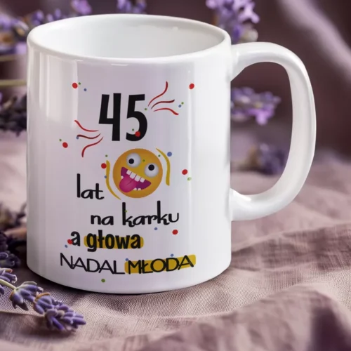 Biały ceramiczny kubek z kolorowym napisem 45 lat na karku a głowa nadal młoda, otoczony kwiatami lawendy.