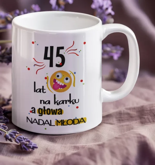 Biały ceramiczny kubek z kolorowym napisem 45 lat na karku a głowa nadal młoda, otoczony kwiatami lawendy.